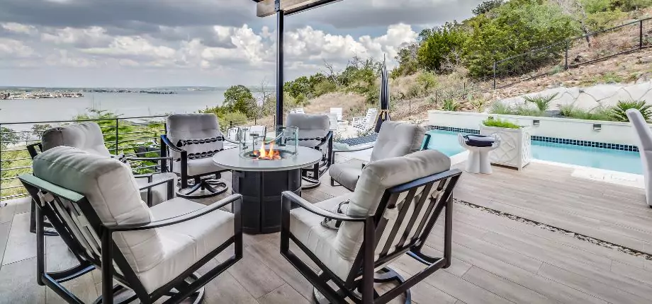 Outdoor patio with cushioned chairs surrounding a fire pit, overlooking a scenic lake view and adjacent to a swimming pool.