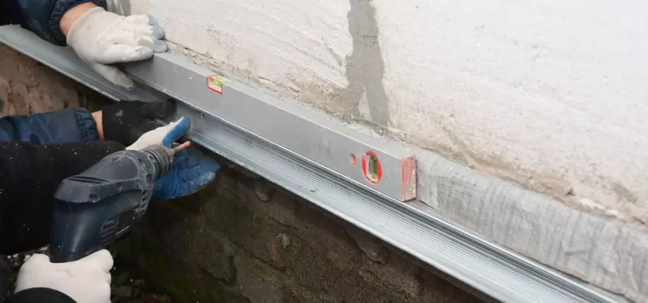Construction worker using a level and drill to align a metal frame on a foundation wall, ensuring precise installation.