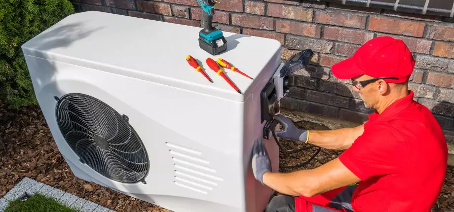 HVAC technician in red uniform performing maintenance on an outdoor air conditioning unit, equipped with tools for repair and installation.