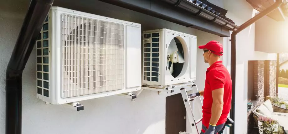 HVAC technician on a ladder inspecting outdoor air conditioning units mounted on a building wall, ensuring optimal cooling performance.