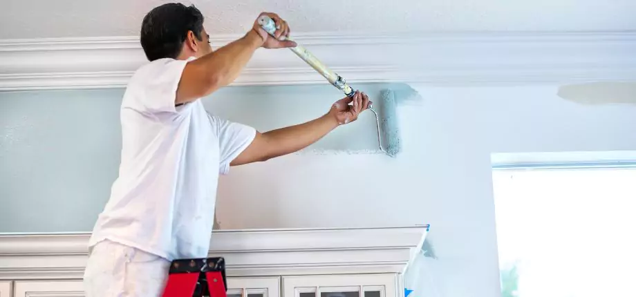 Professional painter on a ladder applying fresh paint to a wall near the ceiling, ensuring a smooth and even finish.