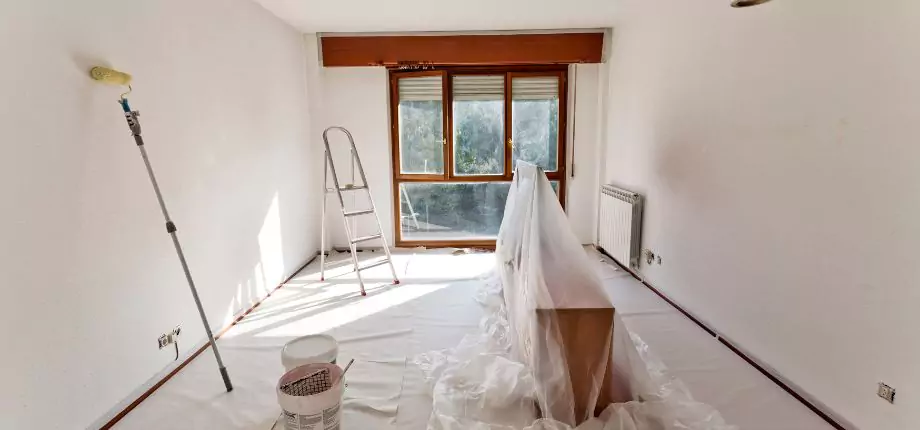 Room prepared for painting with furniture covered in plastic, paint supplies ready, and natural light streaming through large windows.